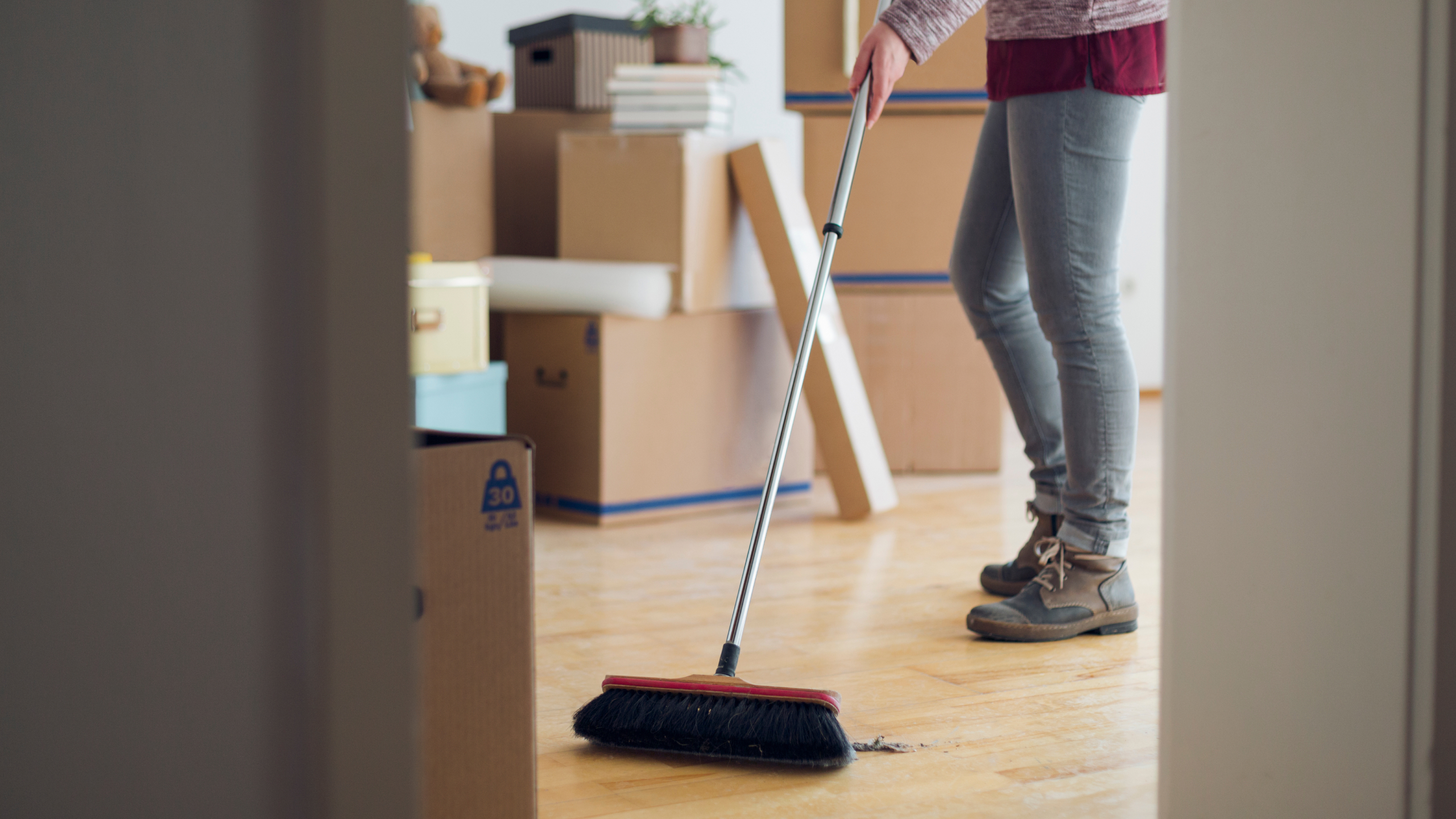 A person sweeping the floor with a broom in a room filled with moving boxes.